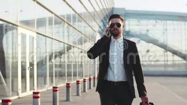 年轻的时尚男士戴着太阳镜在手机上说话，离开机场时拉着手提箱。 成功的生活